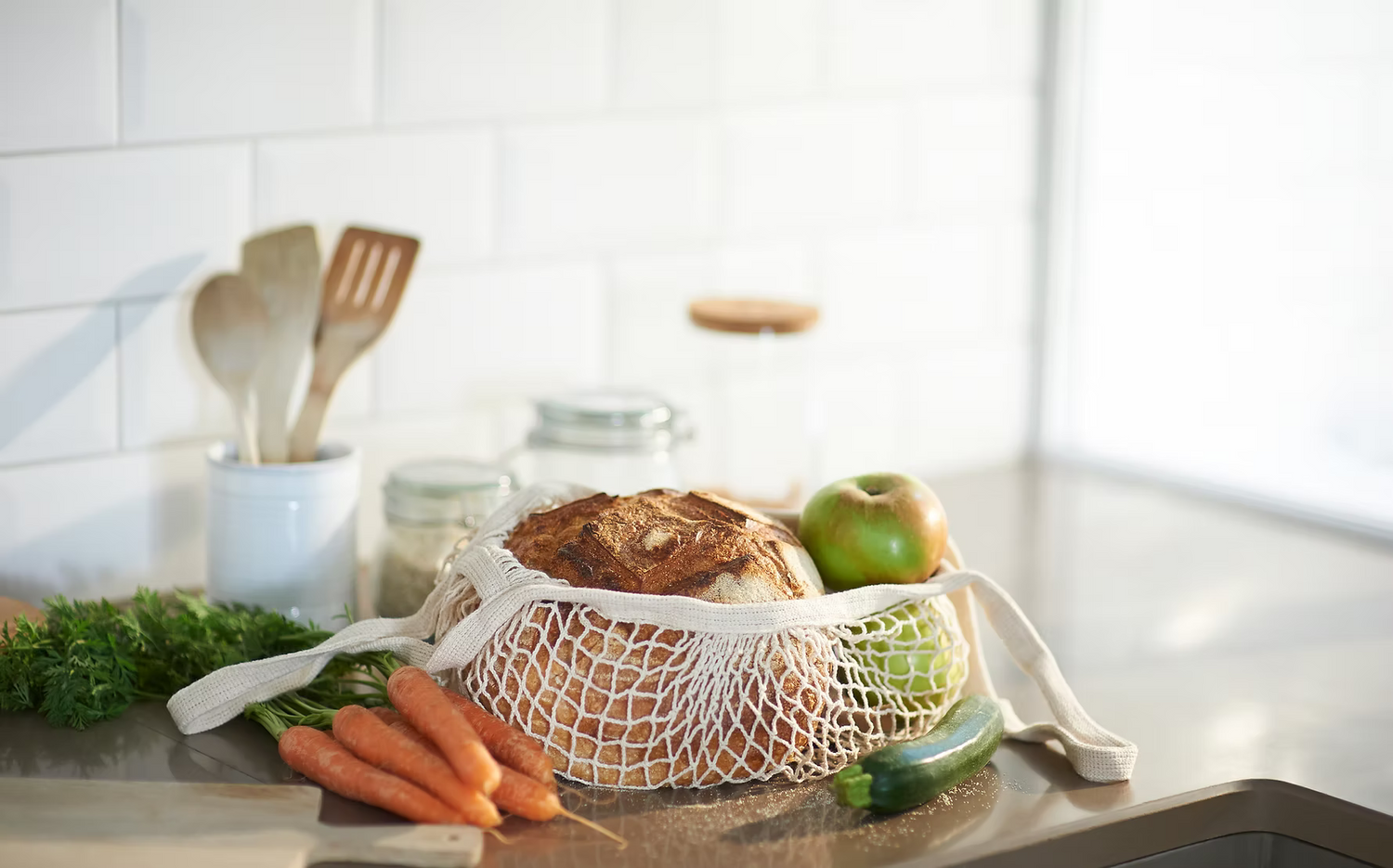 Kitchen counter with a bag of bread, apples, carrots, and cucumbers on a white tiled wall background.
